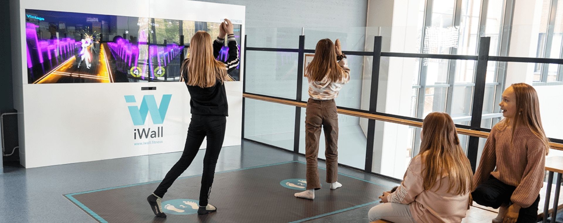 Two girls interact with a dance game on a large screen, while two others watch. The setup is labeled iWall.